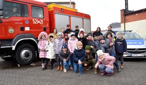 Zdjęcie grupowe na placu apelowym. Policjanci, strażacy oraz dzieci. Po bokach pojazdy służbowe wymienionych służb.