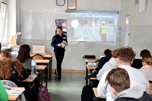 Policjantka w czasie lekcji promuje zawód policjanta. W tle na tablicy plakat zachęcający do wstąpienia w szeregi mundurowych oraz obraz z rzutnika.