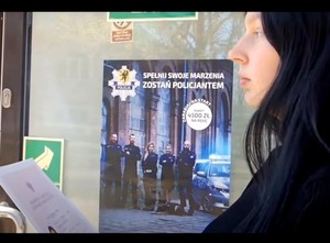 Uczennica trzyma świadectwo ukończenia szkoły. W tle plakat zachęcający do wstąpienia w szeregi policji.