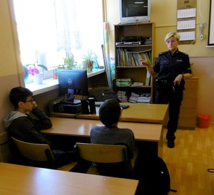 Uczniowie siedzący w ławkach szkolnych. W głębi sali policjantka prowadząca zajęcia.