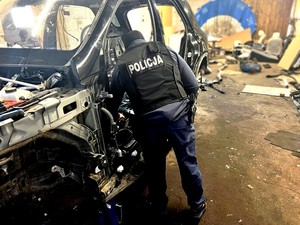 Policjant w kamizelce taktycznej z białym napisem o treści POLICJA prowadzi oględziny stojącego w garażu czarnego mercedesa. Auto jest całkowicie porozkręcane.