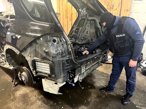 Policjant w kamizelce taktycznej z białym napisem o treści POLICJA prowadzi oględziny stojącego w garażu czarnego mercedesa. Auto jest całkowicie porozkręcane.