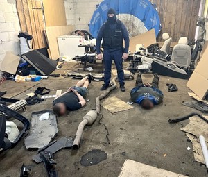 Policjant w kominiarce i kamizelce taktycznej z napisem Policja pilnuje dwóch leżących na ziemi mężczyzn. Zatrzymani leżą na brzuchu i mają założone na rękach kajdanki.