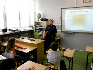 Sala lekcyjna. Dzieci w ławkach. Policjantka prowadzi zajęcia z uczniami.