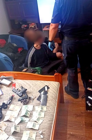 Policjant pilnuje zatrzymanych, którzy siedzą na podłodze z założonymi kajdankami.