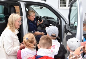 Dzieci stojące przy radiowozie. Policjantka siedzi na miejscu pasażera i opowiada o pracy mundurowych.