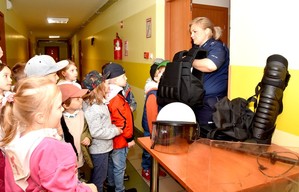 Korytarz komendy. Policjantka prezentuje przedszkolakom sprzęt policyjny. Kask, pałkę, tarczę oraz kamizelkę i ochraniacze na nogi.