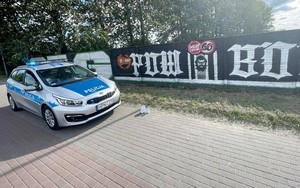Radiowóz policyjny, a przed nim na ziemi puszki z farbą w aerozolu. W tle widoczne grafiti naniesione na kilka przęseł betonowego ogrodzenia.