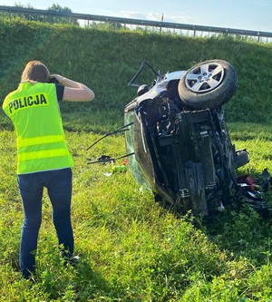 Technik kryminalistyki wykonuje zdjęcie rozbitego auta. Policjant w żółtej kamizelce odblaskowej z czarnym napisem Policja. Na drugim planie rozbite auto leżące na prawym boku.