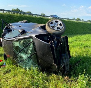 Leżąca na prawym boku rozbita kia. na drugim planie widać nasyp autostrady, na którym zaparkowany jest policyjny radiowóz. Pogoda słoneczna. Dobra widoczność.