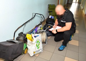 Policjant prowadzi na korytarzu komendy oględziny odzyskanych przedmiotów. Na zdjęciu widoczna miedzy innymi kosiarka spalinowa, wąż do podlewania oraz rozkładany fotel ogrodowy.