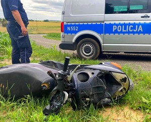 Po lewej stronie stoi umundurowany policjant. Na pierwszym planie widoczny jest przewrócony  w kukurydzy motocykl, a za nim radiowóz policyjny.