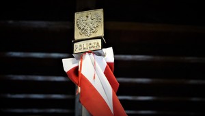 Biało-czerwona szarfa na ciemnym tle, a powyżej orzeł RP oraz napis policja.