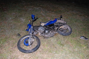 Motocykl przewrócony na prawym boku, leżący na trawie. przy tylnym kole leży męski but. Zdjęcie wykonano po zapadnięciu zmroku.