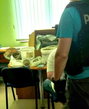 Policjant w kamizelce taktycznej z napisem policja stoi przy siedzącym na krześle zatrzymanym. Na drugim planie widoczny karton i wiadra z zabezpieczonymi narkotykami. Zdjęcie wykonano w pomieszczeniu biurowym na tle zielonych żaluzji okiennych.