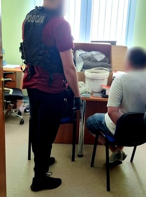 Policjant w kamizelce taktycznej z napisem policja stoi przy siedzącym na krześle zatrzymanym. Na drugim planie widoczny karton i wiadra z zabezpieczonymi narkotykami. Zdjęcie wykonano w pomieszczeniu biurowym na tle zielonych żaluzji okiennych.