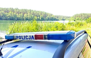 Dach policyjnego radiowozu z kogutem i napisem Policja na tle jeziora i przybrzeżnej trzciny.