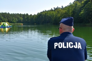 Policjant patrzy w stronę jeziora. Na plecach widoczny biały napis o treści Policja.