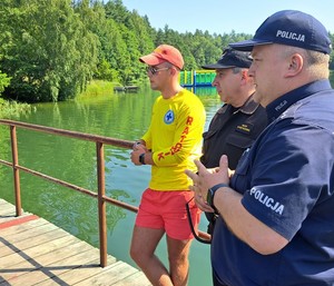 Policjant, strażak i ratownik stoją na pomoście patrząc w stronę kąpieliska. W tle jezioro i las.