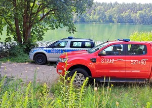 Czerwony auto straży pożarnej, a za nim oznakowany radiowóz policyjny. Zdjęcie wykonano patrząc z boku pojazdów na tle jeziora, trzciny i lasu.