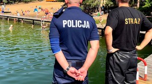 Policjant i strażak stoją na pomoście i patrzą na kąpielisko w jeziorze.