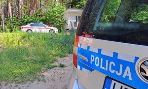 Tył policyjnego radiowozu na tle białego auta osobowego straży pożarnej. Auta stoją na dzikiej plaży, w tle widoczny las.