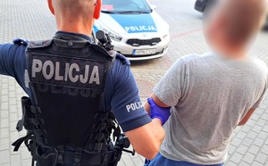 Policjant prowadzi zatrzymanego mężczyznę w kierunku radiowozu.