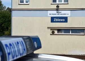 W lewej dolnej części zdjęcia lekko rozmazany kogut policyjny z napisem policja, a w centralnej części zdjęcia budynek PKP z dwiema tablicami. Na pierwszej koloru niebieskiego, biały napis o treści Zblewo. Nad tą tablicą widnieje kolejna biała z niebieskim napisem o treści: PKP Polskie Linie Kolejowe S.A.