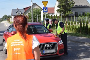 Na pierwszym planie zwrócona plecami kobieta w pomarańczowej kamizelce odblaskowej z białym napisem na plecach o treści: Jestem ambasadorem bezpieczeństwa - szlaban na ryzyko. W tle policjantka rozmawia z kierowcą kontrolowanego audi. W pobliżu stoi funkcjonariusz ochrony kolei w żółtej kamizelce odblaskowej.