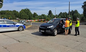 Służby biorące udział w akcji rozmawiają z kierowcą. Po lewej stronie radiowóz policyjny, a w tle otwarty przejazd kolejowy.