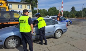 Służby biorące udział w akcji rozmawiają z kierowcą. Po lewej stronie radiowóz policyjny, a w tle otwarty przejazd kolejowy.