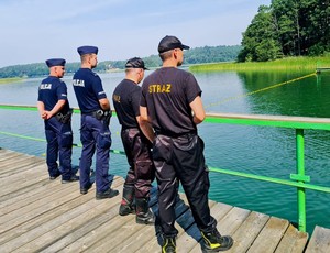 Policjanci i strażacy stoją na pomoście. W oddali widać trzcinę, drzewa i drugi brzeg jeziora.