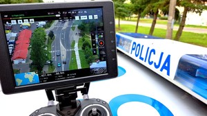Policjant trzyma w ręku urządzenie do sterowania dronem. Na urządzeniu zamontowany jest monitor na którym widoczny jest obraz z kamery.