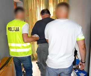 Policjanci prowadzą zatrzymanego mężczyznę. Jeden z f-szy ma założone kamizelkę odblaskową z napisem policja, a drugi opaskę na ręku z napisem Policja.