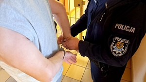 Policjant zakłada podejrzanemu kajdanki na ręce z tyłu. Na lewym ramieniu policjanta widoczna tarcza z wyszytym logiem starogardzkiej komendy.