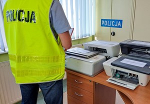 Policjant w żółtej kamizelce odblaskowej prowadzi w biurze oględziny zabezpieczonego sprzętu komputerowego. W tle szafa pancerna z białym napisem policja na granatowym tle.