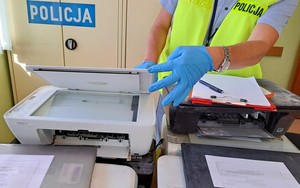 Policjant w żółtej kamizelce odblaskowej prowadzi w biurze oględziny zabezpieczonego sprzętu komputerowego. W tle szafa pancerna z białym napisem policja na granatowym tle.