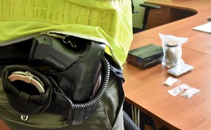 Policjant prowadzi w biurze oględziny zabezpieczonych narkotyków. Funkcjonariusz ma założoną żółtą kamizelkę. Na pierwszym planie przypięta do pasa służbowego broń i kajdanki, a na drugim planie leżą na biurku zabezpieczone środki.