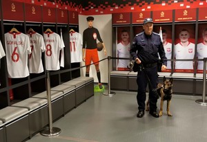 Policjant z psem w pomieszczeniu. Na wieszakach wiszą koszulki sportowe polskiej reprezentacji piłki nożnej.