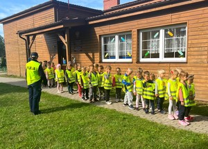 Dzieci ustawiają się do wyjścia. Policjantka oraz przedszkolaki mają założone żółte kamizelki odblaskowe. W tle budynek przedszkola.