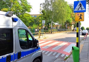 Policyjny radiowóz przed oznakowanym przejściem dla pieszych. Po prawej stronie widoczny znak informacyjny: przejście dla pieszych oraz poniżej tablica informacyjna tak zwana Agatka, która informuje kierowców o tym, że przejście dla pieszych jest szczególnie uczęszczane przez dzieci.
