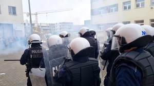 Policyjna grupa szybkiego reagowania z tarczami i w kaskach zmierza w stronę agresywnej grupy osób.