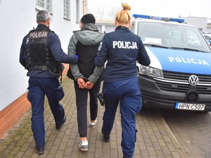 Policjanci prowadzą zatrzymanego. Mężczyzna ma założone na rękach trzymanych z tyłu kajdanki policyjne. Policjanci są w umundurowaniu służbowym.