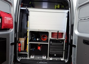 Nowy ambulans pogotowia ruchu drogowego. Otwarte skrzydła drzwiowe z tyłu volkswagena. widoczne wyposażenie pojazdu min. słupki drogowe, kolczatka oraz apteczka. Pozostała część wyposażenia znajduje się za zamkniętą roletą.