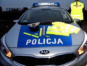 Zaparkowany oznakowany radiowóz policyjny poza obszarem zabudowanym. Na masce samochodu czapka funkcjonariusza ruchu drogowego oraz kamizelki i elementy odblaskowe. W górnej części zdjęcia na dachu radiowozu widoczna zapalona galeria z napisem Policja.