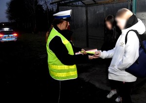 Policjantka wręcza odblaski osobie oczekującej na przystanku autobusowym.