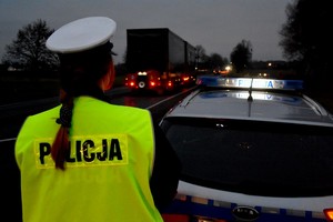 Policjantka w żółtej kamizelce odblaskowej z napisem policja stoi przy oznakowanym radiowozie. W pobliżu znajduje się droga o dużym natężeniu ruchu.