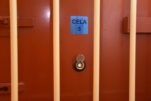 Żółte kraty, a za nimi bordowe drzwi z tabliczką, na której widnieje napis cela nr 5.