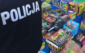 Po lewej stronie widoczne plecy policjantka (kurtka służbowa z białym napisem POLICJA), a w prawej rozłożone na stoisku sklepowym fajerwerki i petardy.