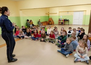 Policjantka prowadzi zajęcia z dziećmi na sali gimnastycznej. Dzieci siedzą w grupie na podłodze  i słuchają wypowiedzi funkcjonariuszki.
